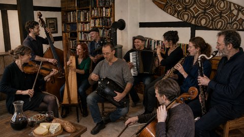 A group of musicians playing various instruments in a cozy room. Bread and wine are on the table.