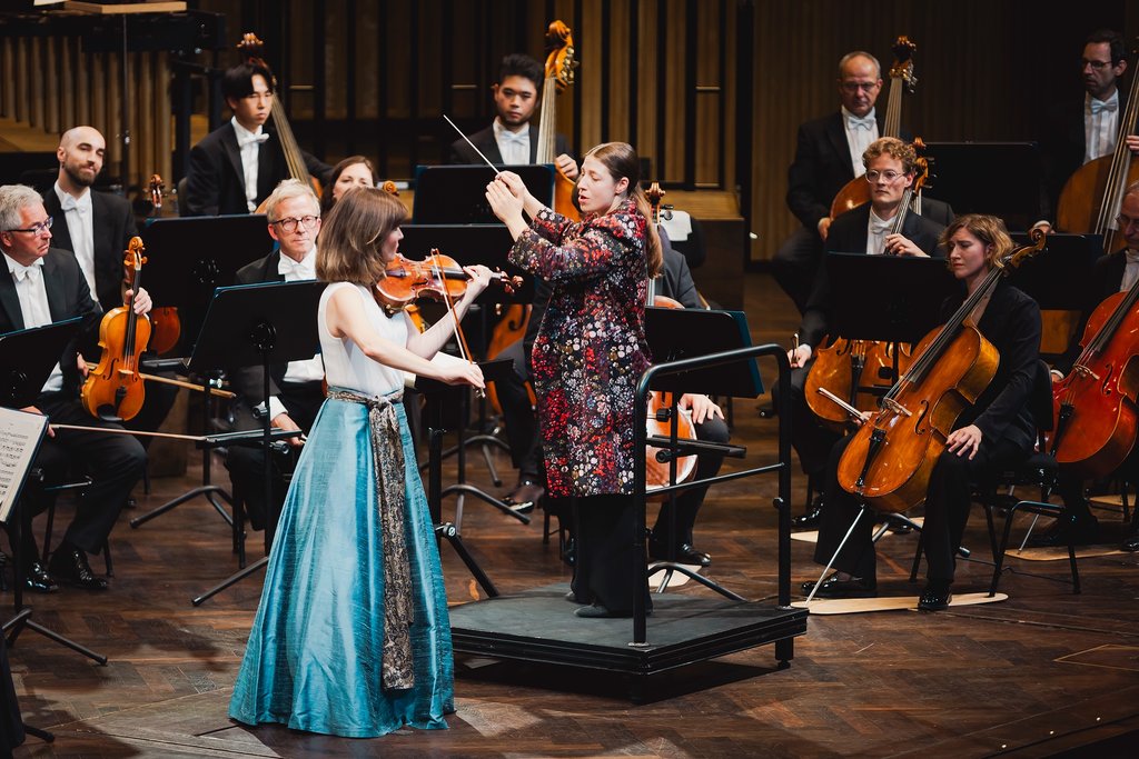Eine Dirigentin leitet ein Orchester, während eine Geigerin in einem blauen Kleid auf der Bühne spielt.