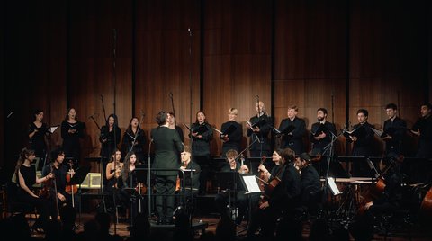 Ein Orchester und Chor treten auf einer Bühne auf. Die Musiker und Sänger stehen vor einem braunen Hintergrund.