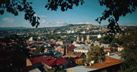 Ein Panorama von Tiflis, Georgien, mit historischen Gebäuden und Hügeln im Hintergrund, gesehen durch Bäume im Vordergrund.