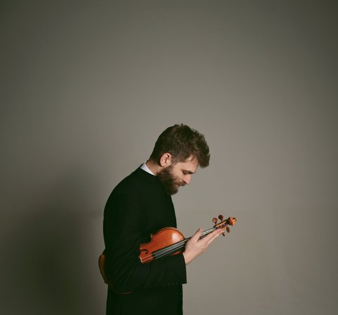 Ein Mann in schwarzem Anzug hält eine Geige und schaut nachdenklich auf das Instrument, vor neutralem Hintergrund.