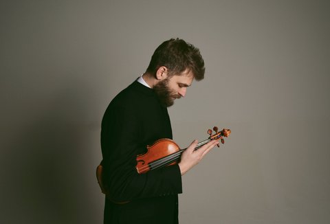Ein Mann in schwarzem Anzug hält eine Geige und schaut nachdenklich auf das Instrument, vor neutralem Hintergrund.