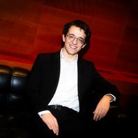 A person in formal attire sits relaxed on a black leather sofa against a warmly lit, wooden backdrop.