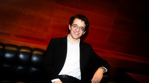 A person in formal attire sits relaxed on a black leather sofa against a warmly lit, wooden backdrop.