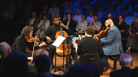 Ein Streichquartett aus vier Männern sitzt in einem Kreis in der Mitte eines Konzertsaales und spielt gemeinsam.