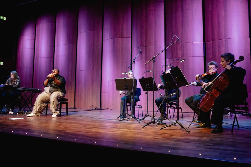 Eine Band spielt auf einer Bühne mit lila Hintergrundbeleuchtung. Musiker spielen Cello, Violinen und ein Keyboard.