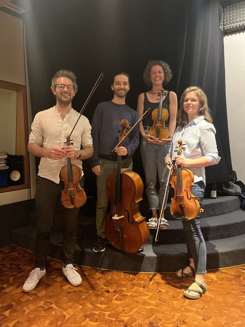 Vier Musiker mit Streichinstrumenten posieren auf einer Bühne. Zwei halten Geigen, einer eine Bratsche und einer ein Cello.