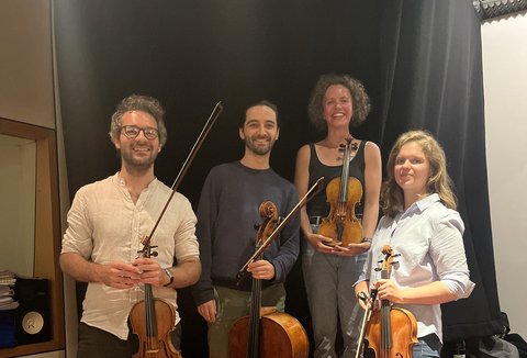 Vier Musiker mit Streichinstrumenten posieren auf einer Bühne. Zwei halten Geigen, einer eine Bratsche und einer ein Cello.