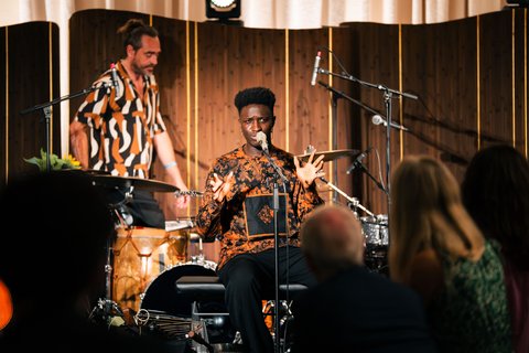 Ein Musiker sitzt vor einem Mikrofon in einem Club und spielt ein Instrument, während ein Schlagzeuger ihn begleitet.