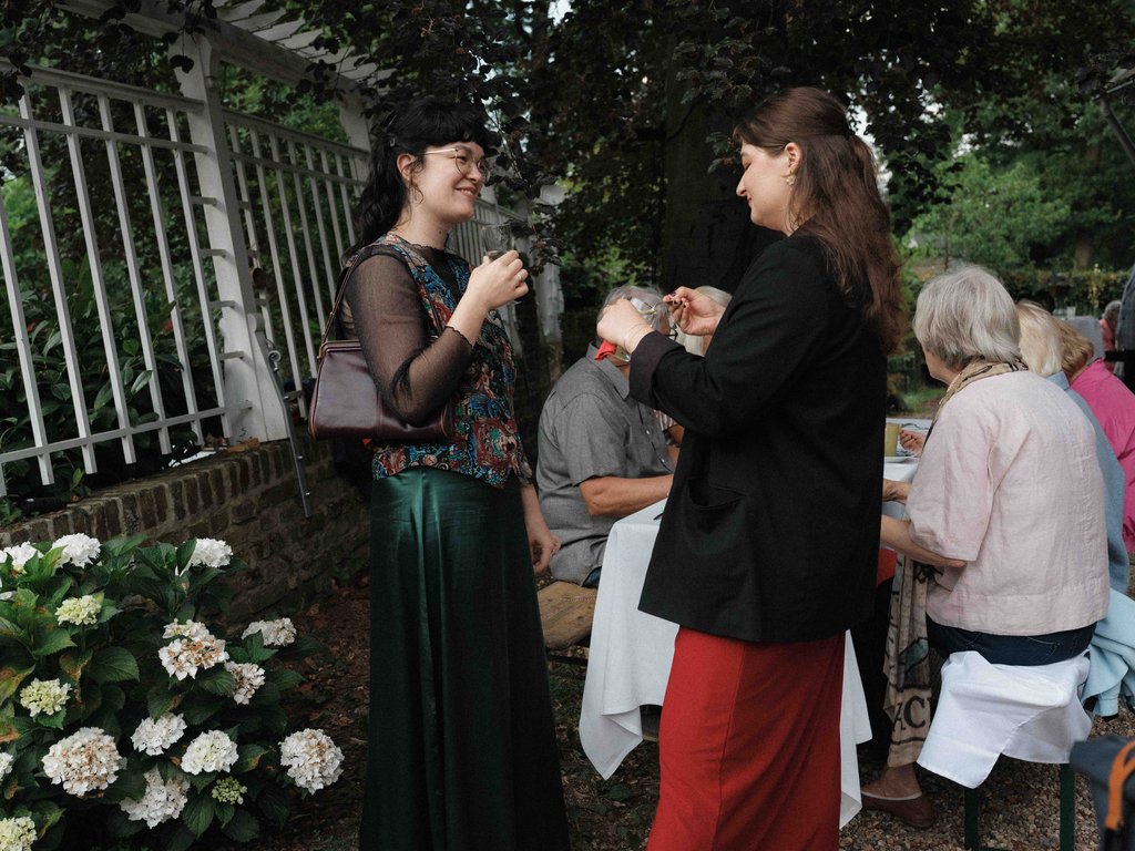 Zwei Frauen unterhalten sich bei einer Gartenparty. Im Hintergrund sitzen andere Gäste an einem Tisch. Blumen schmücken die Szene.