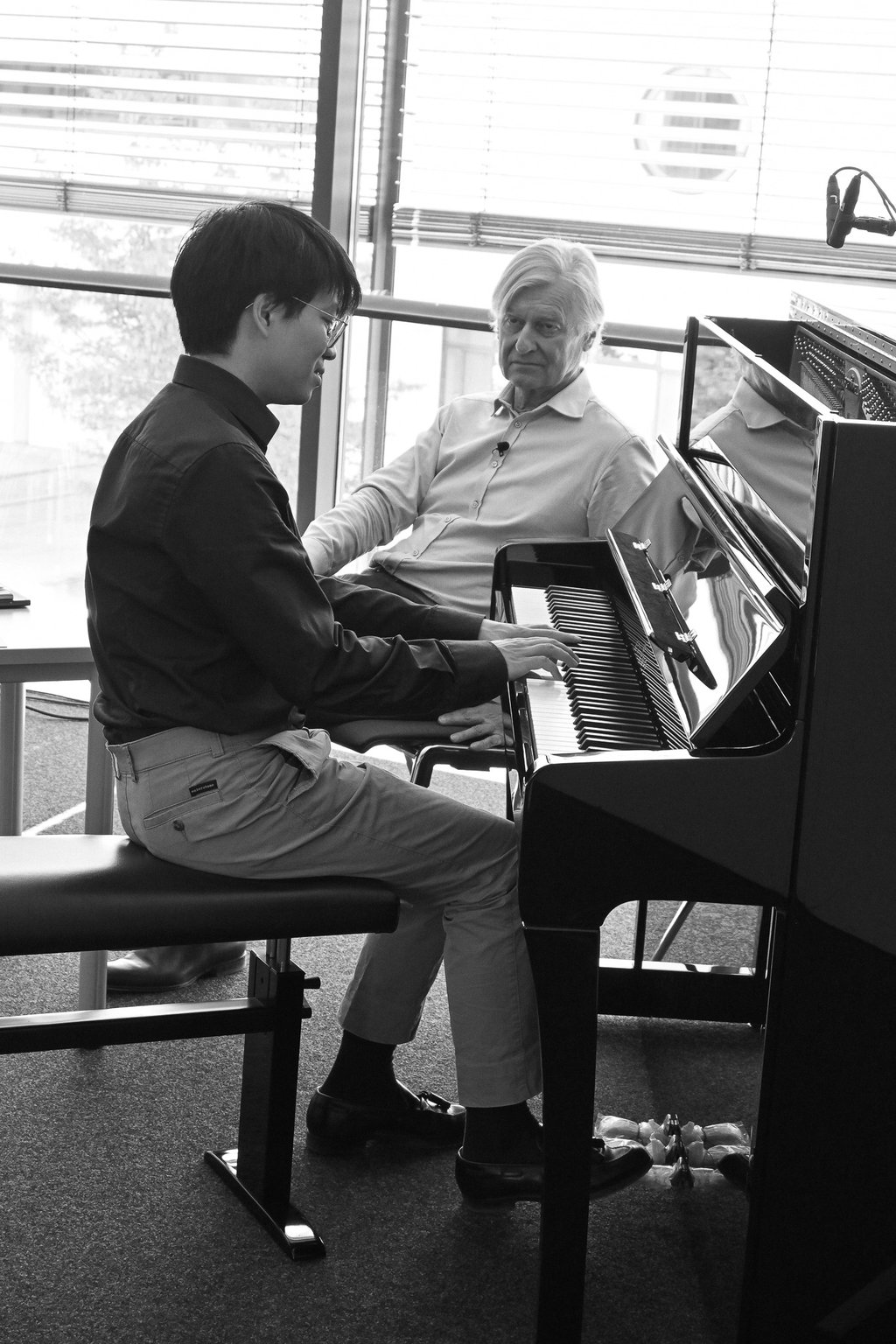 Zwei Personen an einem Klavier, eine spielt, während die andere aufmerksam zuhört. Schwarzweißfoto in einem modernen Raum.