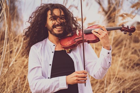 A smiling man with long, curly hair plays the violin in a natural setting with reeds and grass in the background.