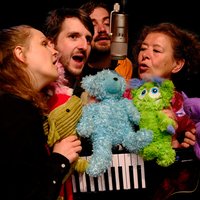 Fünf Personen singen in ein Mikrofon, umgeben von bunten Plüschtieren. Die fröhliche Gruppe wirkt in einem musikalischen Auftritt.