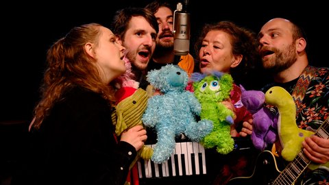 Fünf Personen singen in ein Mikrofon, umgeben von bunten Plüschtieren. Die fröhliche Gruppe wirkt in einem musikalischen Auftritt.