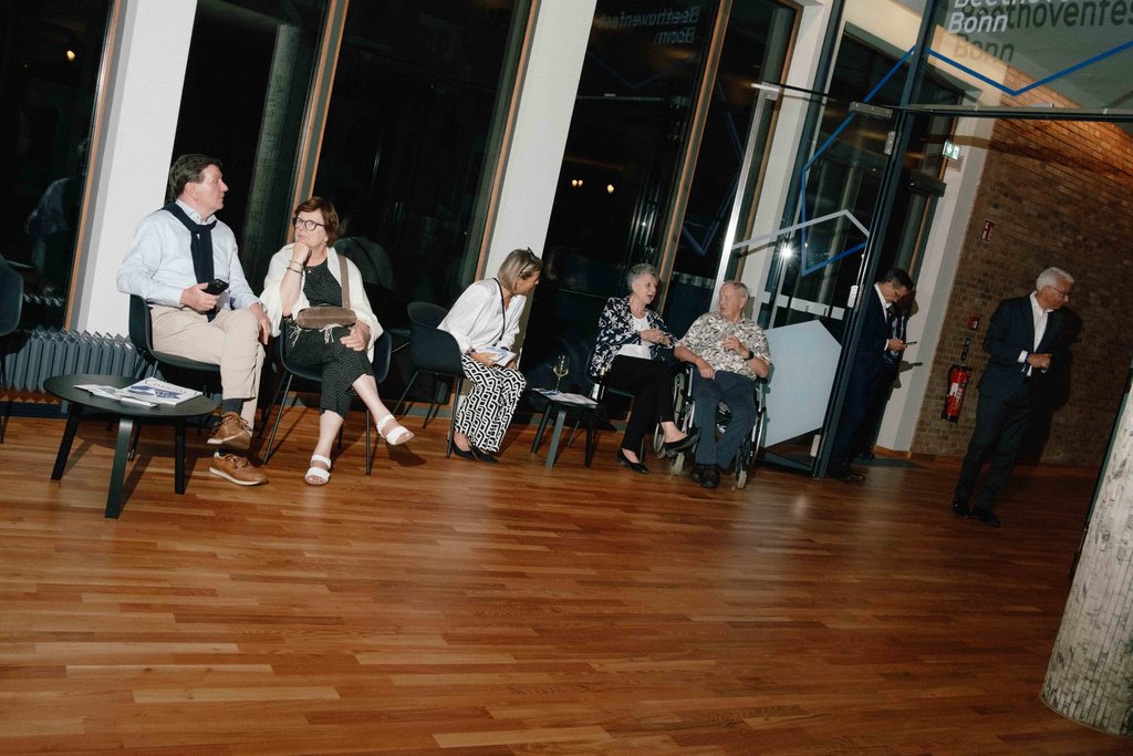Mehrere Menschen sitzen und unterhalten sich in einem eleganten Raum mit Holzboden und großen Fenstern. Eine entspannte Atmosphäre herrscht vor.
