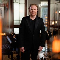 A man in a black suit stands in a stylishly decorated room with soft candlelight in the foreground.