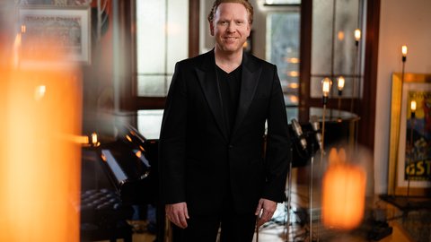 A man in a black suit stands in a stylishly decorated room with soft candlelight in the foreground.