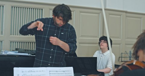 Ein Dirigent leitet eine Probe, während ein Pianist im Hintergrund spielt. Musiknoten sind im Vordergrund zu sehen.