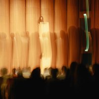 A blurred image of a lecture with a speaker on stage in front of a curtain. Audience members are in the foreground.