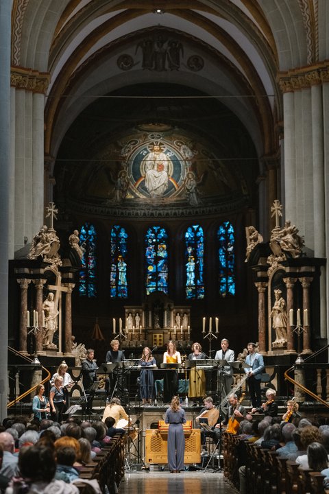 Eine musikalische Darbietung in einer beeindruckenden Kirche mit reich verzierten Altären, Statuen und bunten Glasfenstern. Publikum sitzt andächtig.