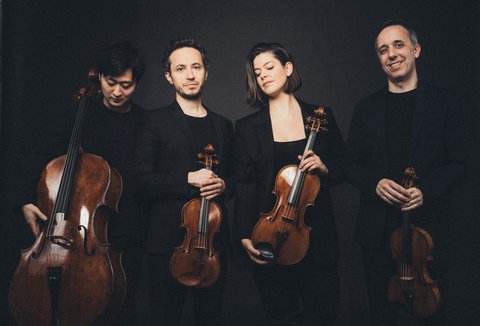 Three men and a woman are standing in a row. They are holding string instruments in front of them. They are all dressed in black and are looking either straight ahead or down, their expressions ranging from serious to smiling. The background is black.