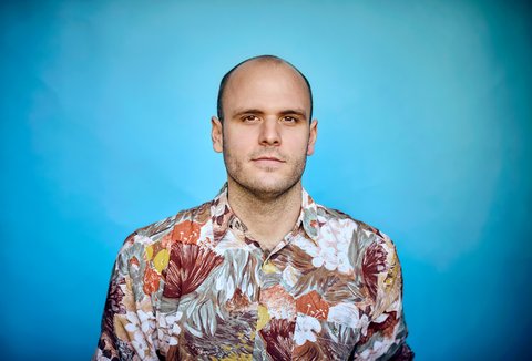 A man is standing in front of a light background. He is wearing a patterned shirt and looking into the camera.