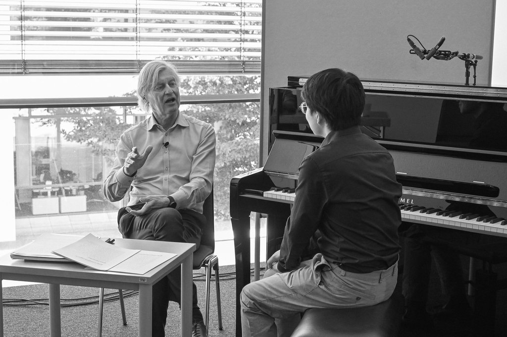 Zwei Männer sitzen in einem Raum mit einem Klavier. Sie unterhalten sich. Auf einem Tisch liegen Notenblätter.