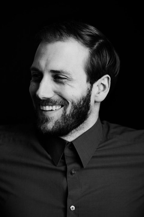 A man is posing in front of a dark backgroud. He is looking away from the camera and laughing.