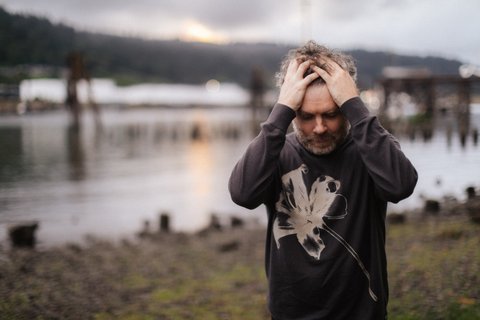 A man is standing in a meadow by a lake; he is wearing a dark jumper with a large white flower printed on it. He has his hands buried in his grey curls and is looking down. The atmosphere is sombre.  © Jason Quigley