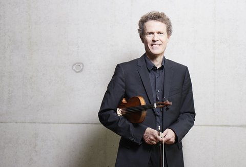 A man in a suit holds a violin and smiles in front of a plain, gray wall.