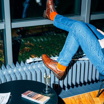 Person mit blauen Jeans und orangenen Socken, Füße auf der Fensterbank, Glas Sekt und Bücher auf dem Tisch. Man sieht nur die Beine.