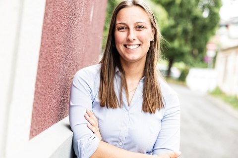 Eine Person in einem hellblauen Hemd lächelt und steht vor einer rot gestrichenen Wand. Im Hintergrund ist ein Weg mit Bäumen zu sehen.