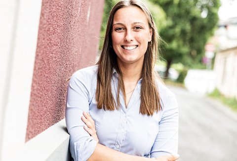 Eine Person in einem hellblauen Hemd lächelt und steht vor einer rot gestrichenen Wand. Im Hintergrund ist ein Weg mit Bäumen zu sehen.
