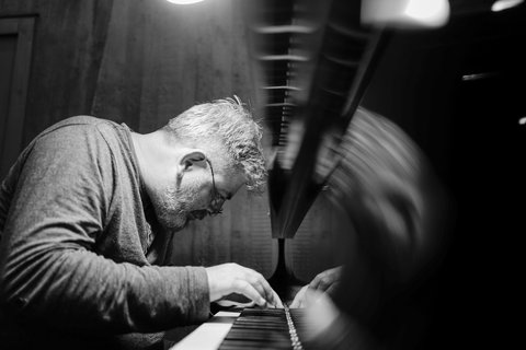 A man with glasses intensely plays the piano. The black-and-white image captures a focused musical performance in a small space.