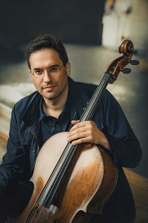 Ein Mann sitzt und hält ein Cello. Er trägt eine dunkle Jacke und blickt freundlich in die Kamera.