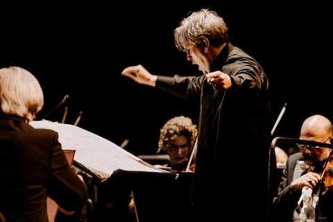 Ein Dirigent leitet ein Orchester mit intensiven Gesten. Musiker spielen konzentriert, während sie die Partitur auf geöffneten Notenständern lesen.