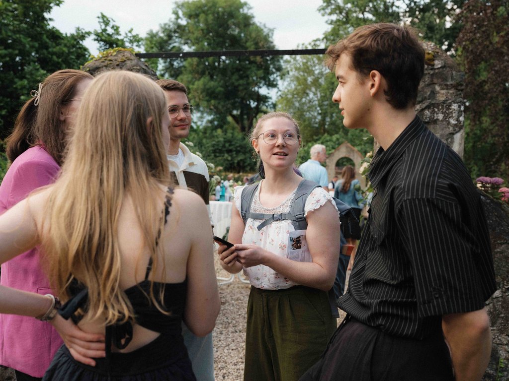 Eine Gruppe junger Menschen unterhält sich auf einer Gartenparty. Sie lachen und genießen die entspannte Atmosphäre im Freien.