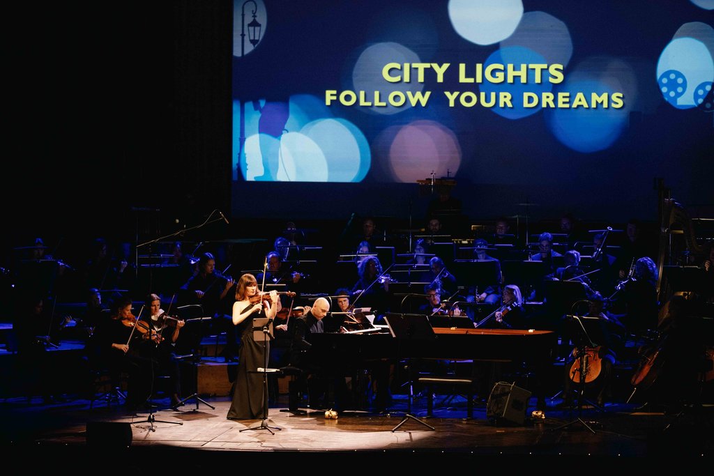 Eine Musikerin singt auf einer beleuchteten Bühne, begleitet von einem Orchester, mit der Aufschrift "City Lights: Follow Your Dreams" im Hintergrund.