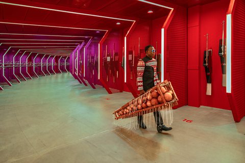 Person in einem futuristischen, roten Gang trägt ein Balafon, ein westafrikanisches hölzernes Xylophon mit runden Resonanzkörpern. Die Beleuchtung ist in violetten und roten Tönen gehalten.