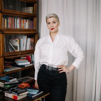 Eine Frau steht vor einem Bücherregal. Sie blickt in die Kamera. Ihre Haare sind kurz und sie trägt eine helle Bluse. Sie stützt den Arm in die Hüfte.