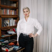 Eine Frau steht vor einem Bücherregal. Sie blickt in die Kamera. Ihre Haare sind kurz und sie trägt eine helle Bluse. Sie stützt den Arm in die Hüfte.