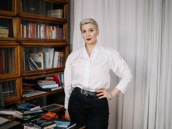Eine Frau steht vor einem Bücherregal. Sie blickt in die Kamera. Ihre Haare sind kurz und sie trägt eine helle Bluse. Sie stützt den Arm in die Hüfte.