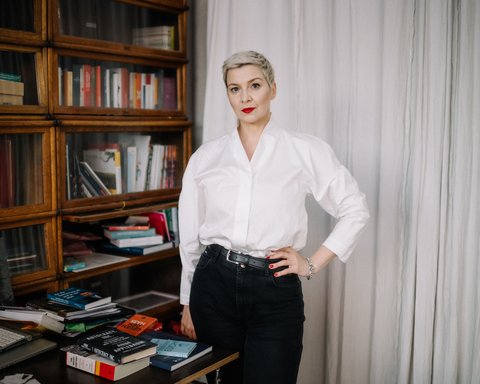 Eine Frau steht vor einem Bücherregal. Sie blickt in die Kamera. Ihre Haare sind kurz und sie trägt eine helle Bluse. Sie stützt den Arm in die Hüfte.