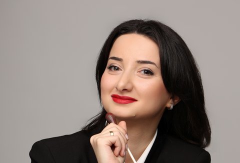 Eine junge Frau mit schwarzen Haare, rotem Lippenstift und schwarzer formeller Kleidung posiert für die Kamera mit einem Baton. Sie lächelt.