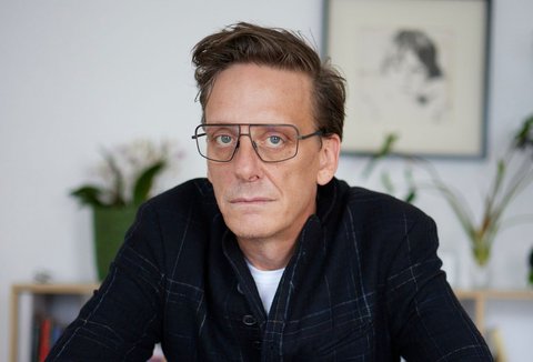 A man is sitting at a table. He has put both arms on the table. He is looking into the camera. He is wearing a dark jacket and glasses.