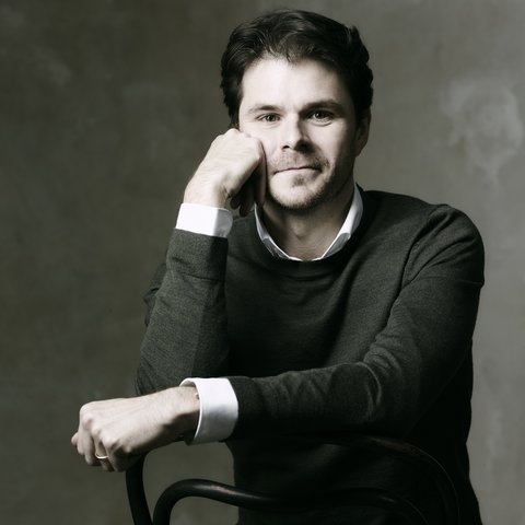 Ein Mann in einem dunklen Pullover sitzt auf einem Stuhl. Er stützt sein Gesicht auf die Hand und lächelt sanft.