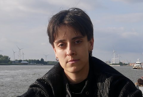 A woman is sitting in front of a river. She is wearing a dark jacket. She is looking into the camera.