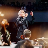 A conductor leads an orchestra, surrounded by soft light, while musicians play with focus and intensity.