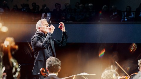 A conductor leads an orchestra, surrounded by soft light, while musicians play with focus and intensity.