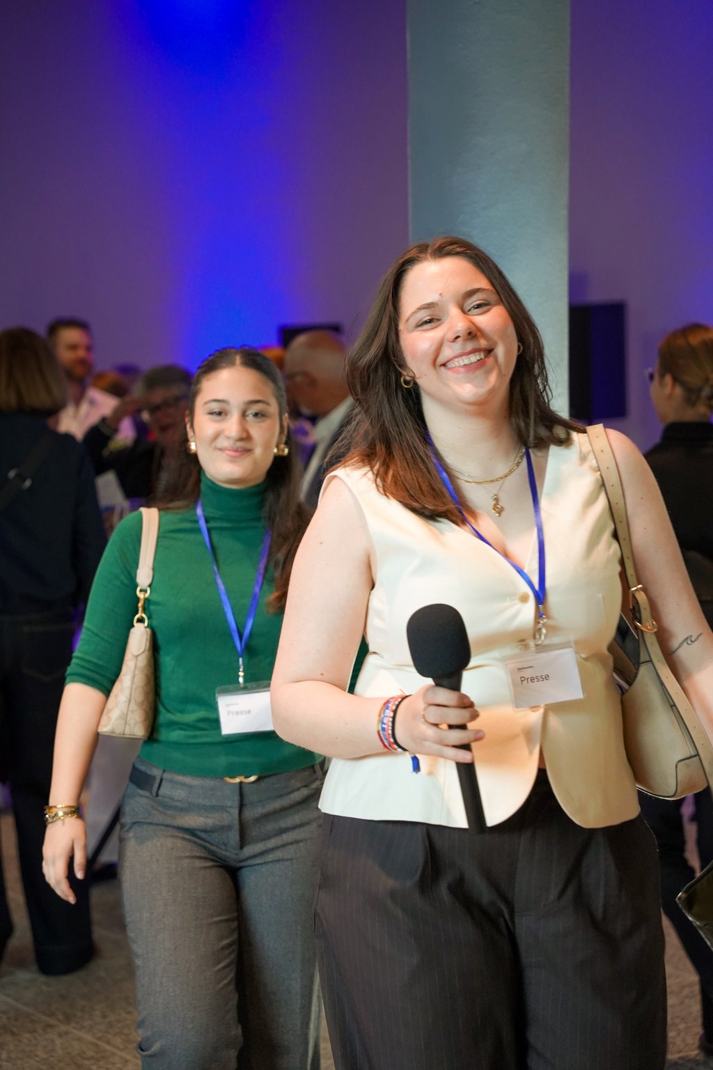 Two women at an event. One holds a microphone, both smiling. Other attendees are visible in the background.